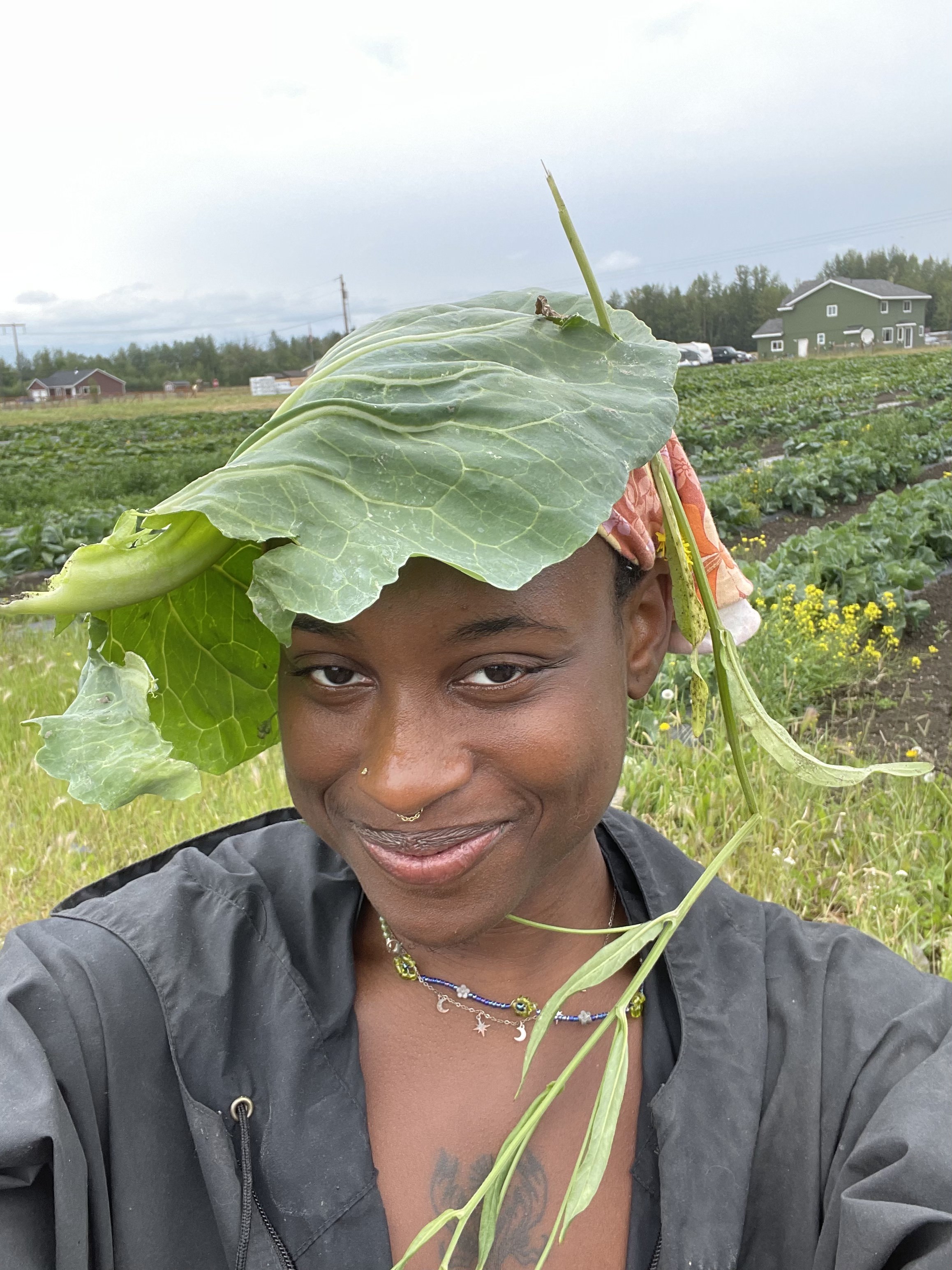 Picture of me with a cabbage leaf on my head. I am smiling and wearing a floral bandana and a black windbreaker.
